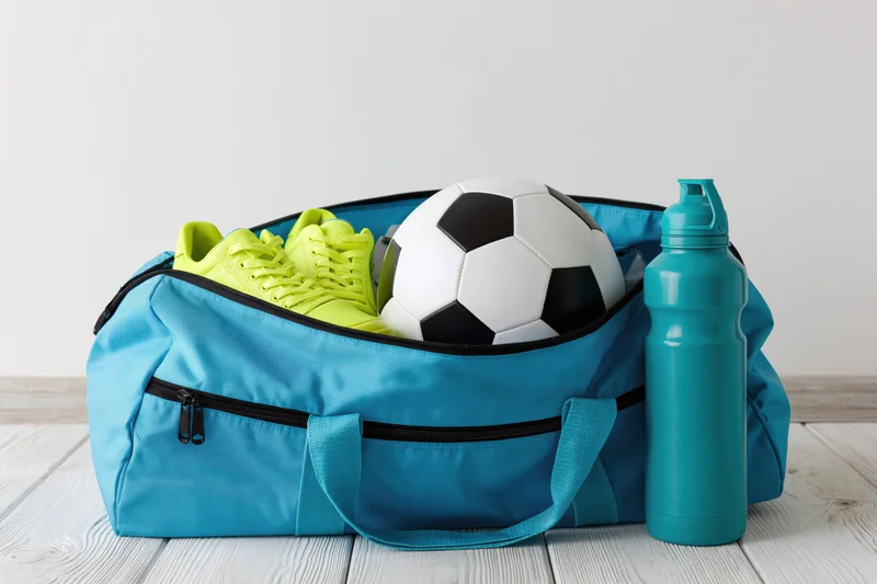 Open cyan sports duffel bag containing soccer ball and lime green cleats with water bottle beside it on wooden floor
