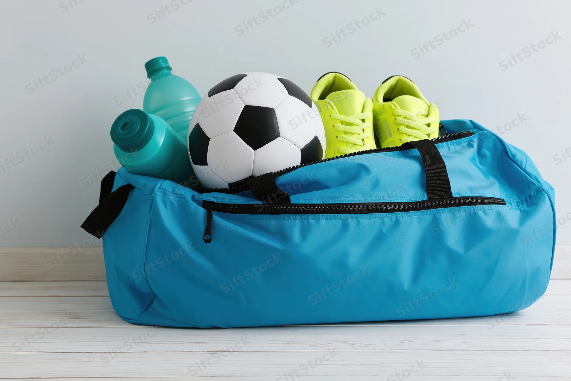 Open cyan sports duffel bag filled with soccer ball, neon yellow cleats and water bottles on wooden floor