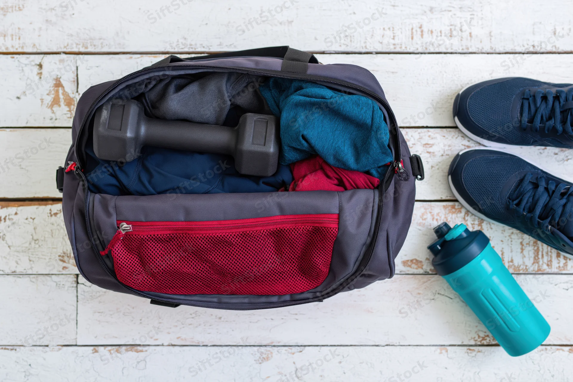 Open gym bag filled with workout clothes and a dumbbell on a wooden floor with sneakers and a water bottle beside it