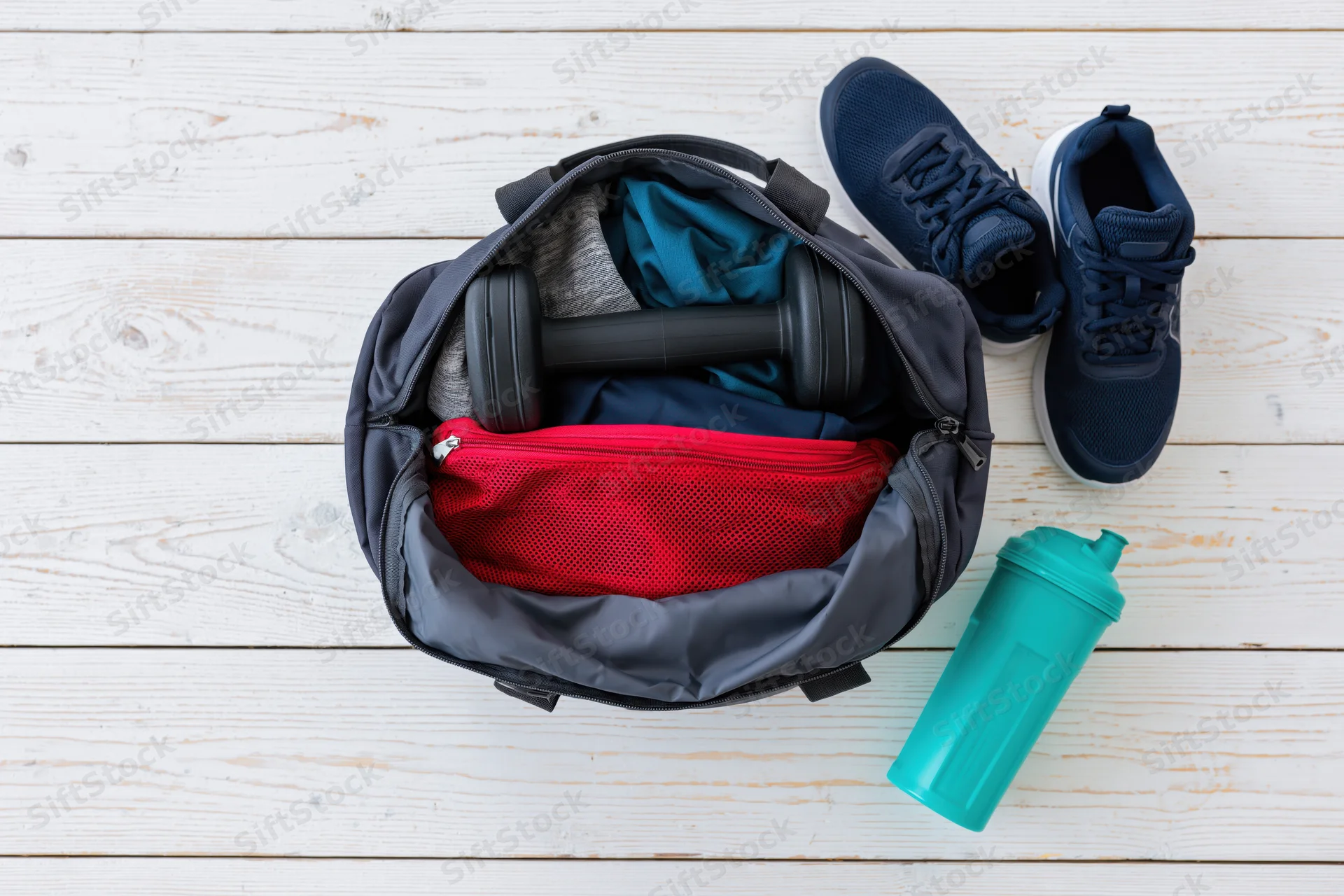 Open gym bag with workout clothes and dumbbell on wooden floor next to sneakers and water bottle