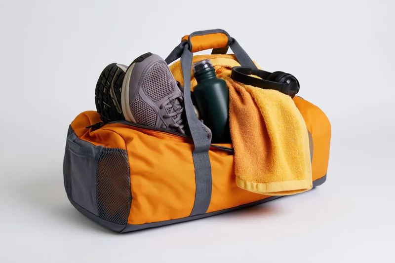 Orange and grey gym duffel bag packed with sneakers, water bottle, towel and headphones on white background