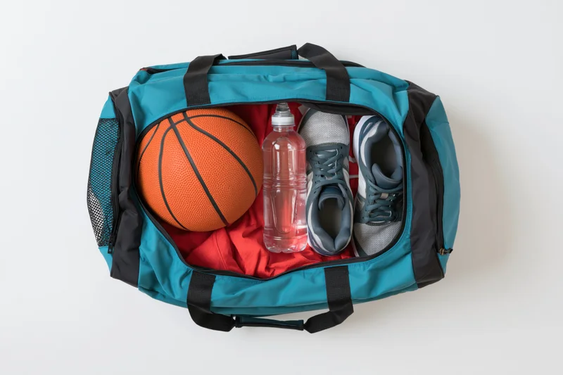 Open teal and black sports duffel bag packed with basketball, sneakers, water bottle, and red shirt on white background