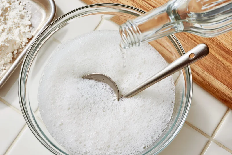 Vinegar being poured into a foamy white batter in a glass bowl on a tiled kitchen counter