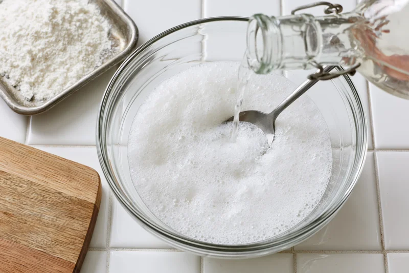Water being poured into a foamy white starter in a glass bowl on a tiled kitchen counter
