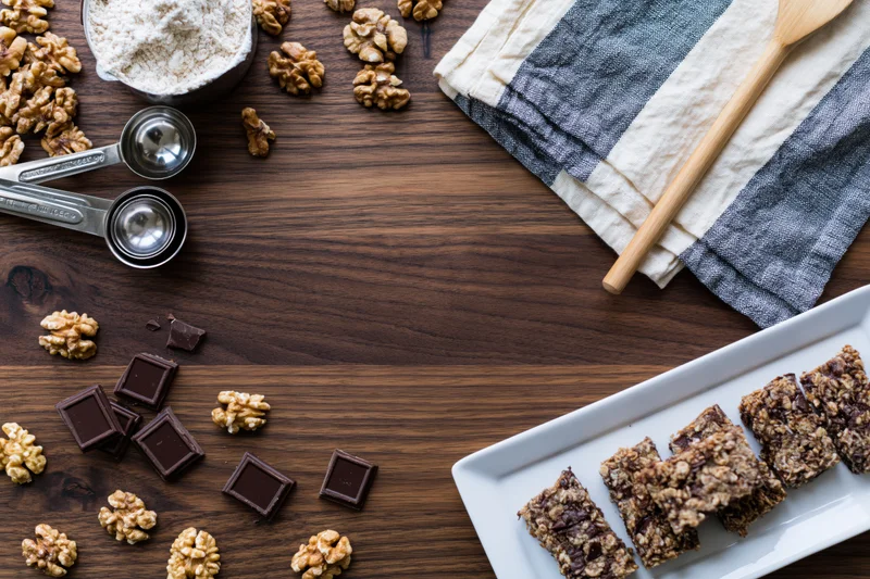 Granola bars with chocolate and walnuts arranged on dark walnut wood table with baking ingredients and utensils