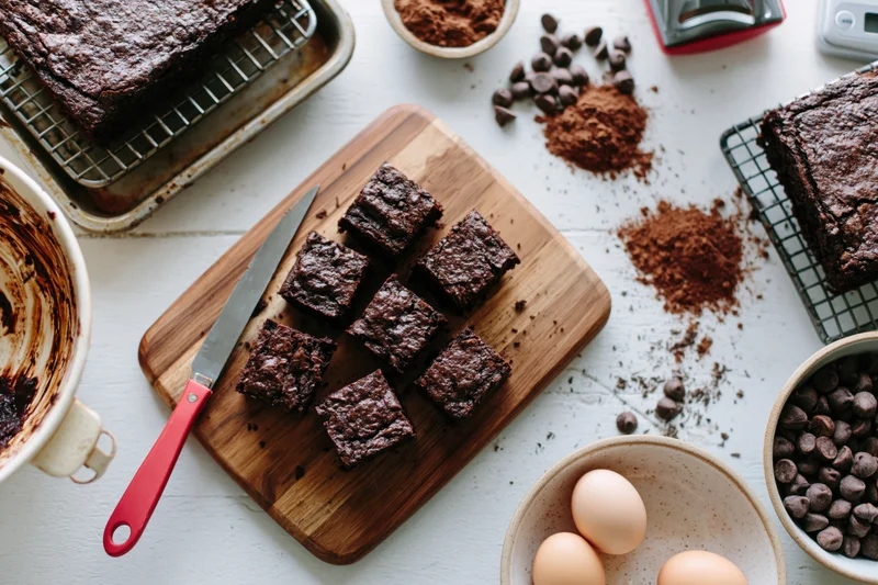 Freshly cut chocolate brownies arranged on a wooden cutting board with baking ingredients around on a white countertop