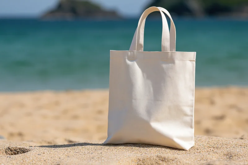 Plain white canvas tote bag standing upright on sandy beach with ocean in background