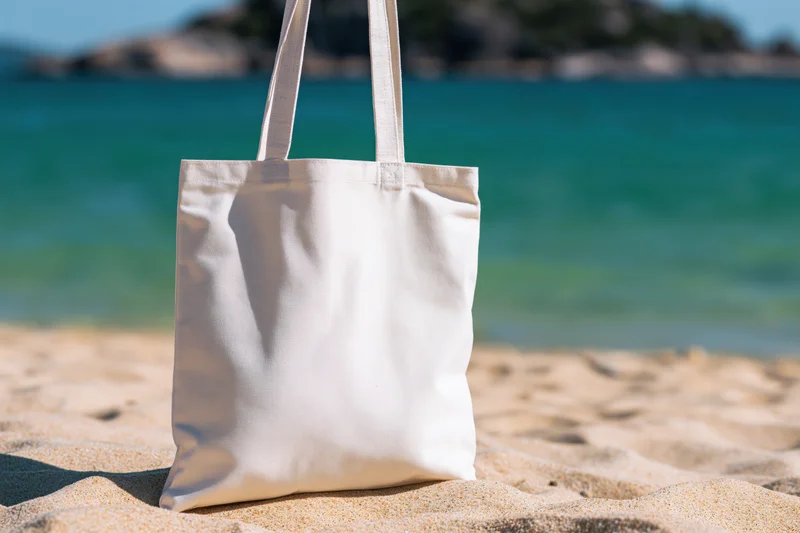 Plain white canvas tote bag standing upright on sandy beach near ocean