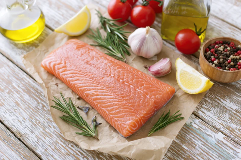 Raw salmon fillet on crumpled parchment paper surrounded by tomatoes, lemon, garlic, rosemary, pepper and olive oil on wooden table