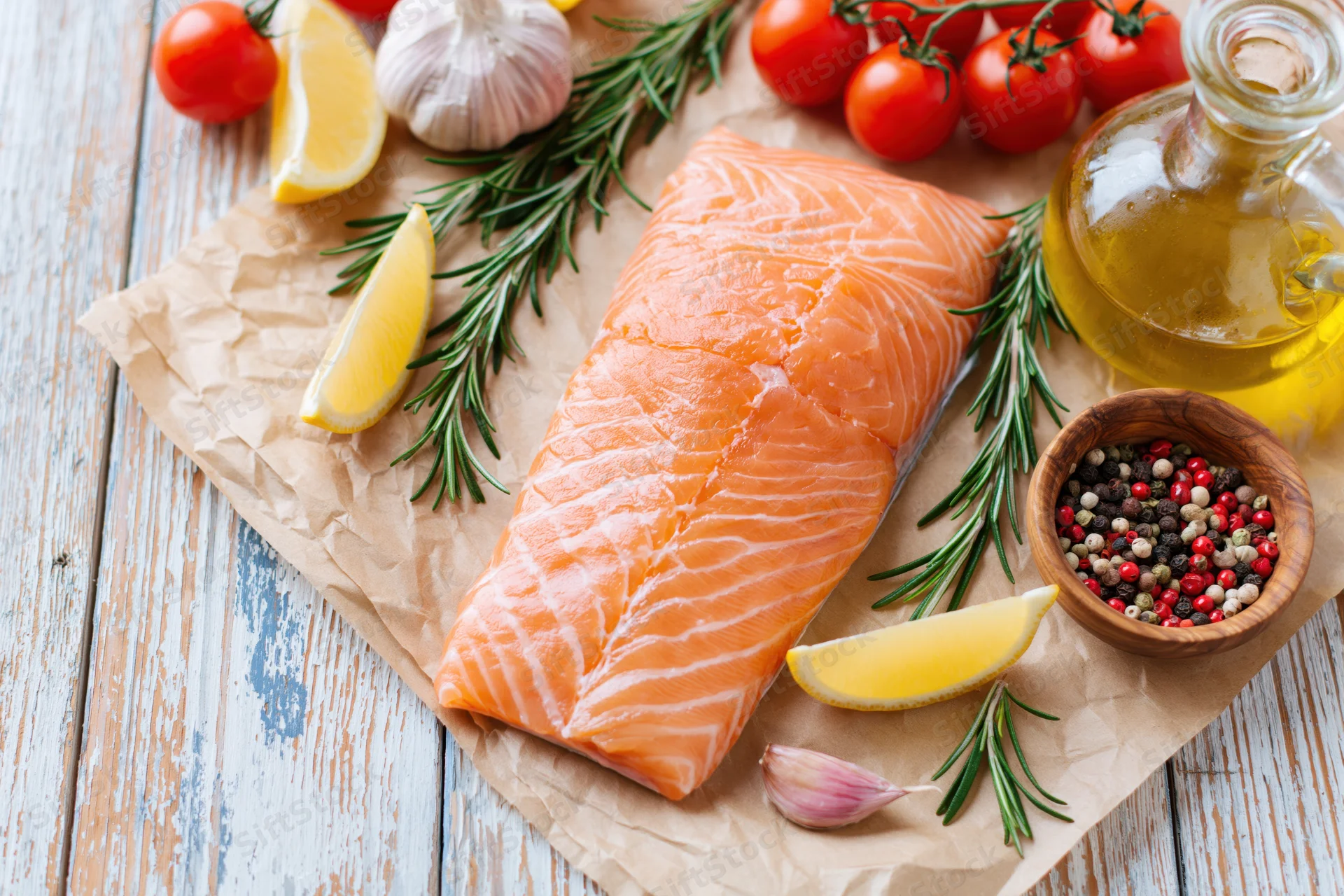Raw salmon fillet on crumpled parchment paper surrounded by tomatoes, lemon, garlic, rosemary, olive oil, and peppercorns on a wooden board