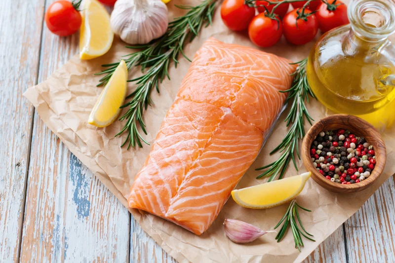 Raw salmon fillet on crumpled parchment paper surrounded by tomatoes, lemon, garlic, rosemary, olive oil, and peppercorns on a wooden board