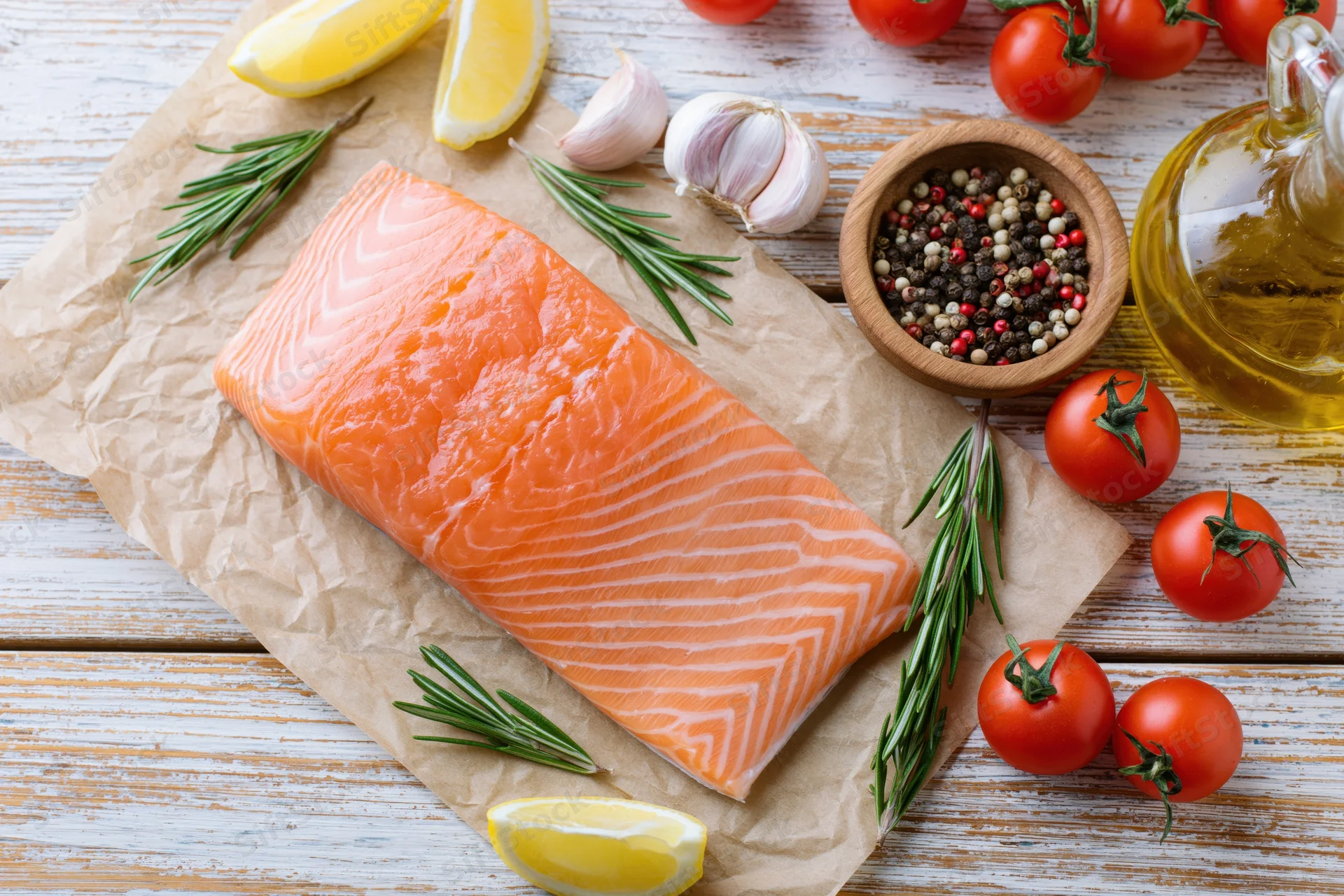 Raw salmon fillet on crumpled parchment paper surrounded by tomatoes, lemon, garlic, rosemary, pepper and olive oil on wooden table