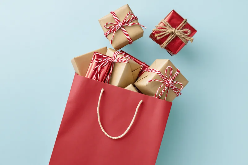 Red paper shopping bag filled with wrapped gift boxes on light blue surface