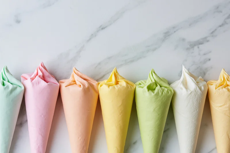 Seven pastel-colored frosting piping bags filled and twisted in a row on a marble surface
