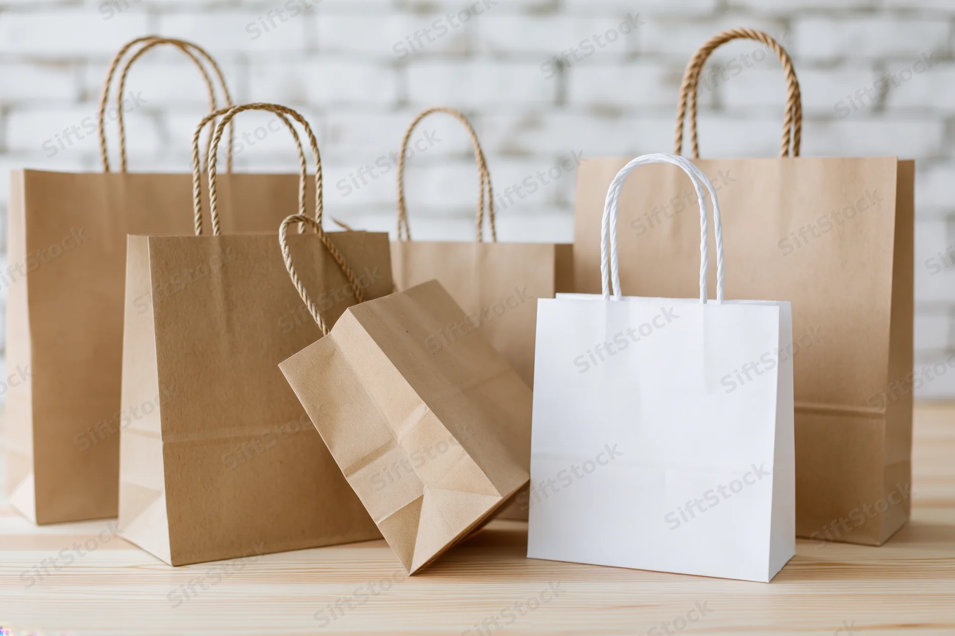 Several kraft paper shopping bags and one white paper bag arranged on a wooden table against a white brick wall