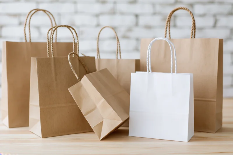 Several kraft paper shopping bags and one white paper bag arranged on a wooden table against a white brick wall