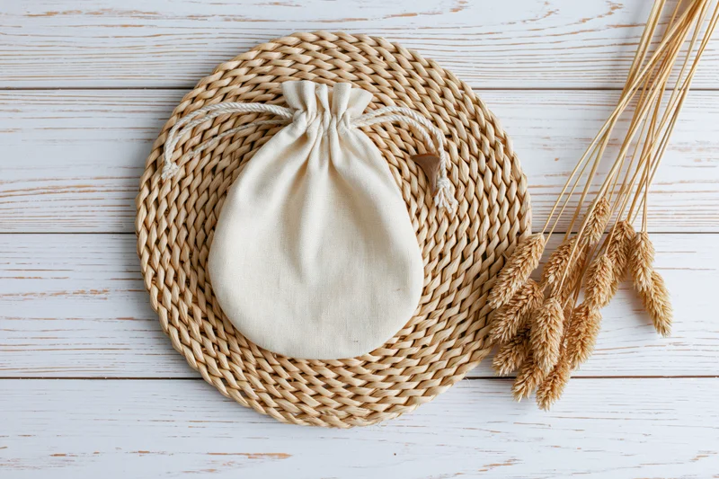 Cream cotton drawstring pouch on woven placemat with dried wheat on white wooden surface