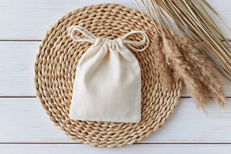 Cream cotton drawstring pouch on round woven mat with dried pampas grass on white wooden surface