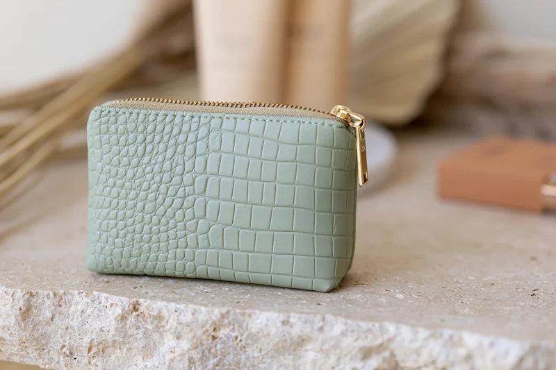 Small sage green crocodile-embossed faux leather cosmetic pouch with gold zipper on stone surface