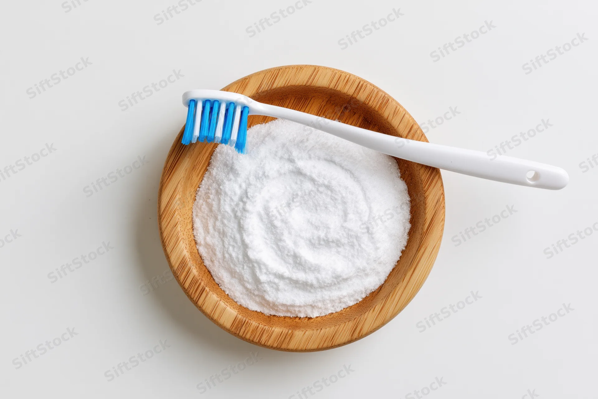 White toothbrush resting on wooden bowl filled with baking soda powder on plain surface