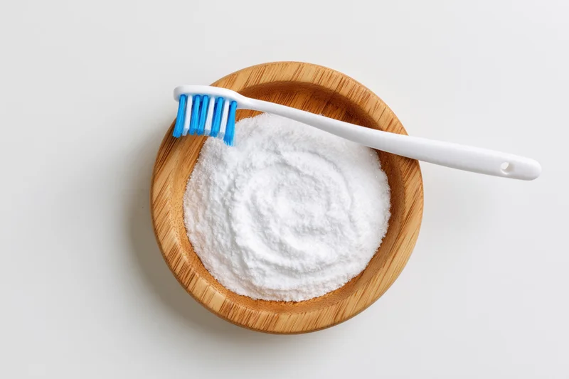 White toothbrush resting on wooden bowl filled with baking soda powder on plain surface