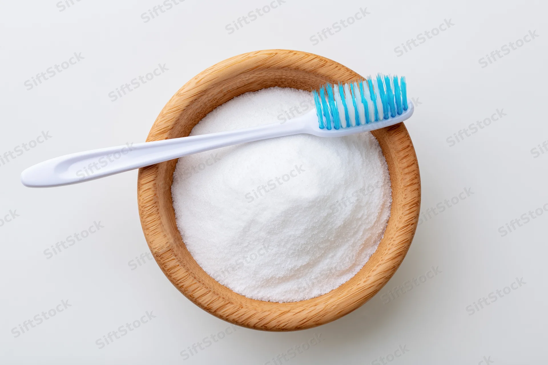 White toothbrush resting on wooden bowl filled with baking soda powder on plain white surface