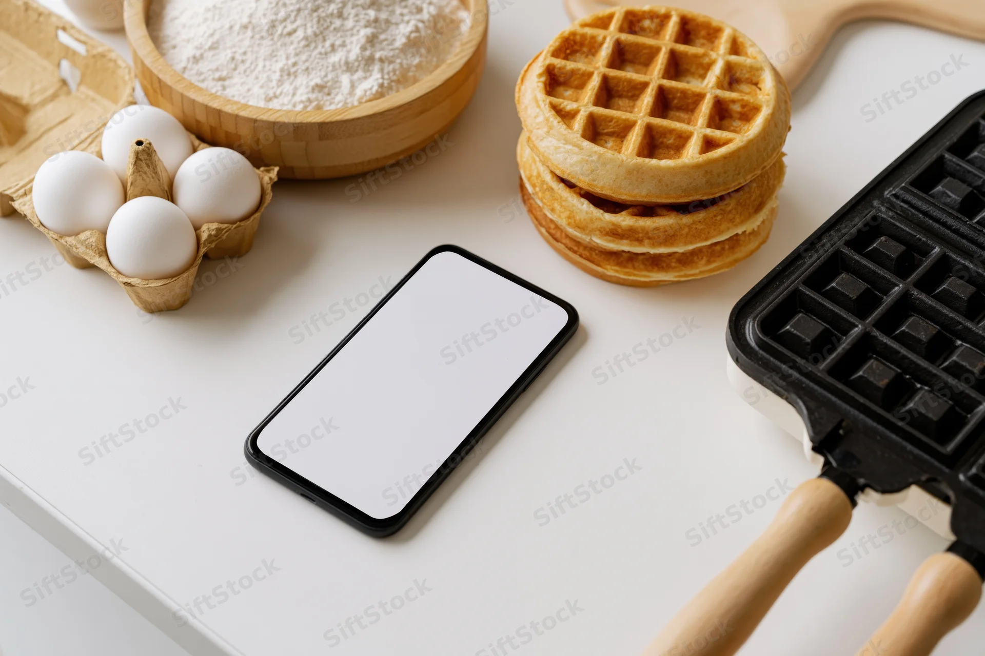 Smartphone with blank screen lying on kitchen counter beside waffles, flour, eggs and waffle iron