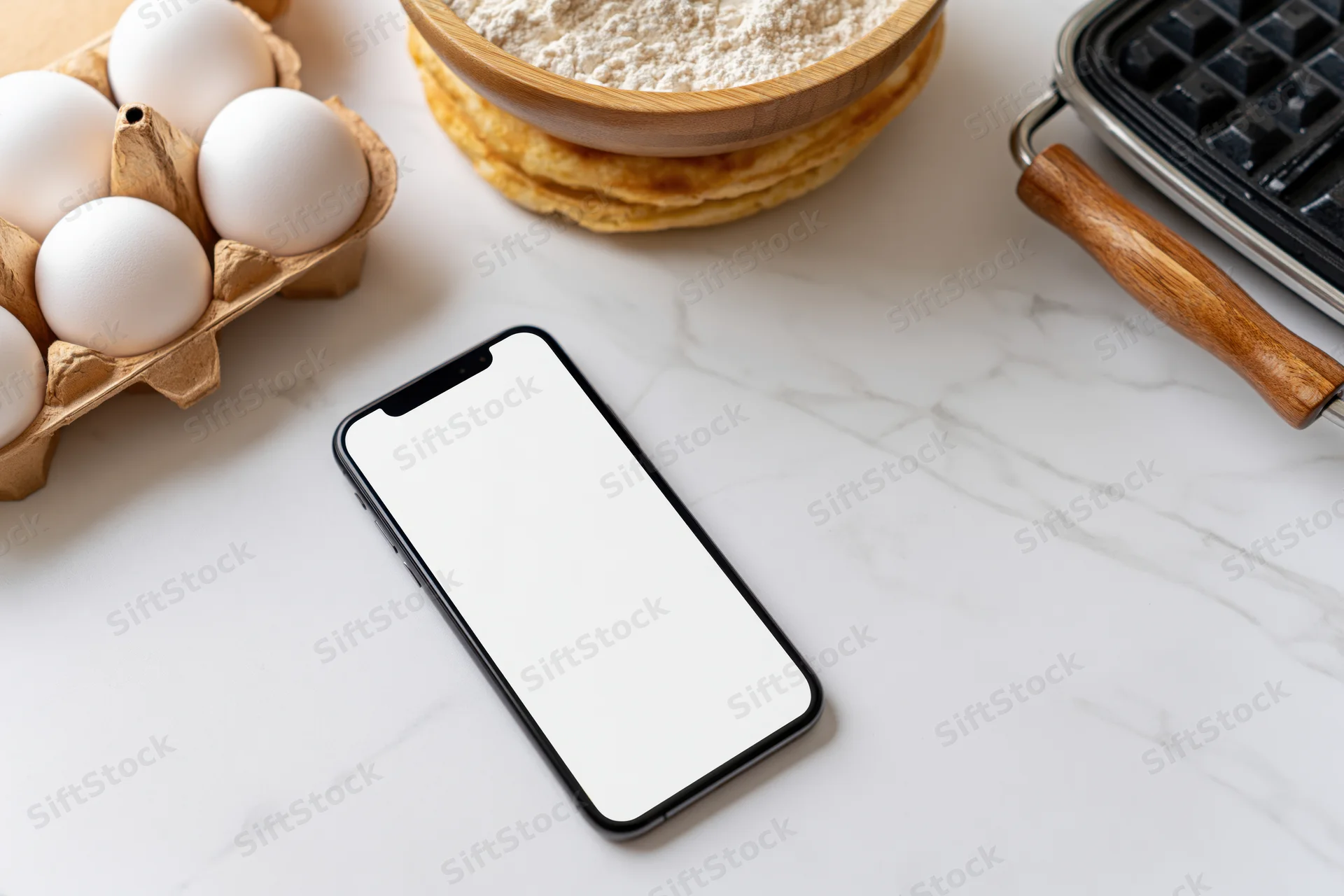 Smartphone with blank white screen lying flat on marble kitchen countertop surrounded by baking ingredients and waffle iron