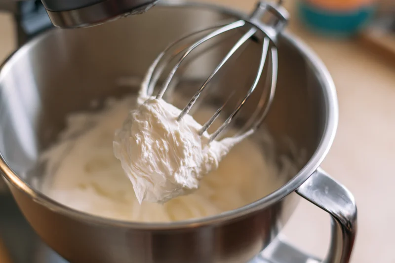 Whisk attachment lifting thick white cream from stainless steel bowl in stand mixer