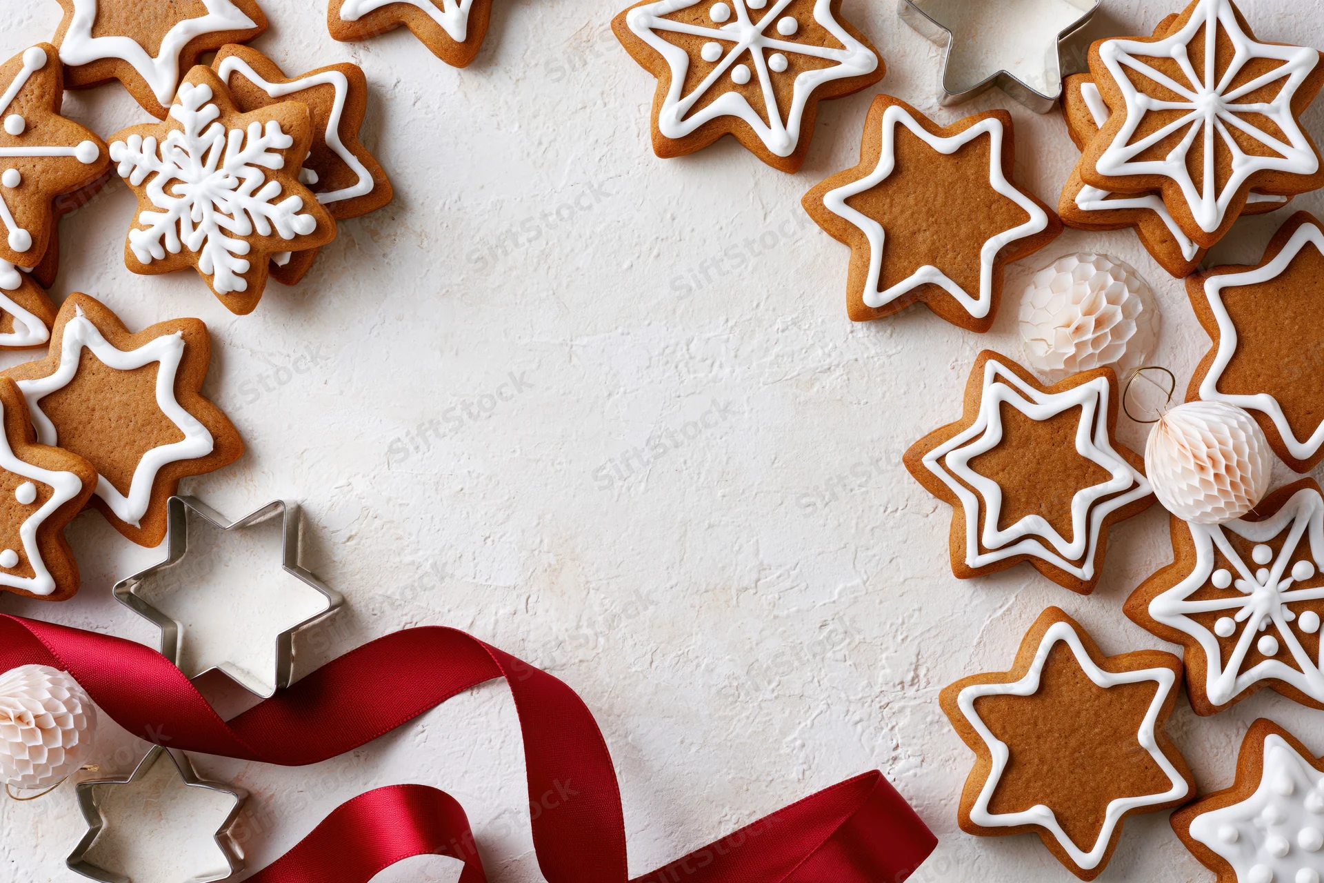 Star-shaped gingerbread cookies with white icing and red ribbon arranged on a white surface with cookie cutters and paper ornaments around the edges