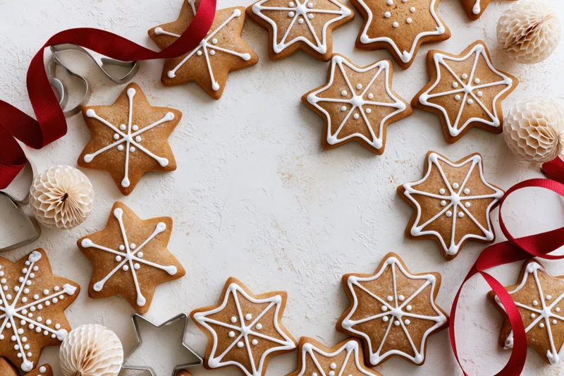 Star-shaped gingerbread cookies with white icing arranged in a circle on a white surface with red ribbons and paper ornaments