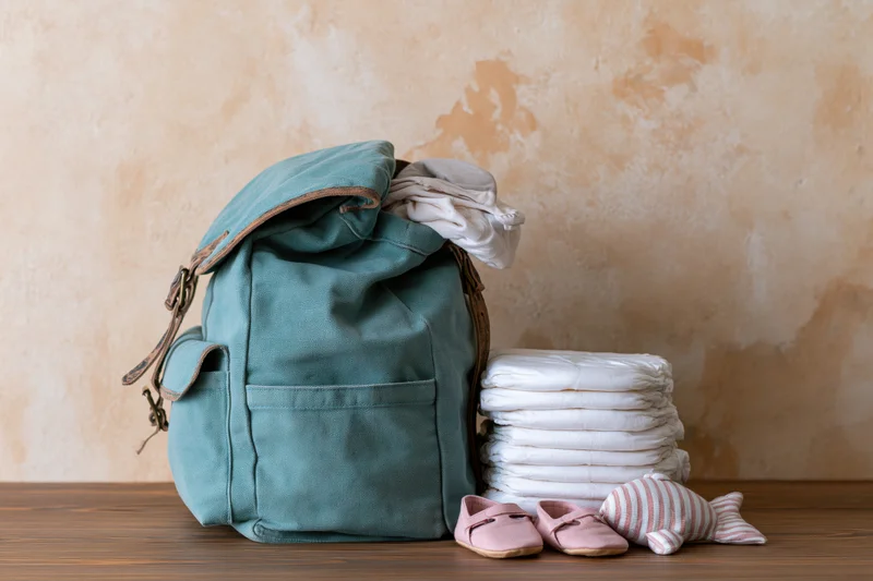 Teal canvas diaper bag backpack with diapers, baby shoes and toy on wooden surface