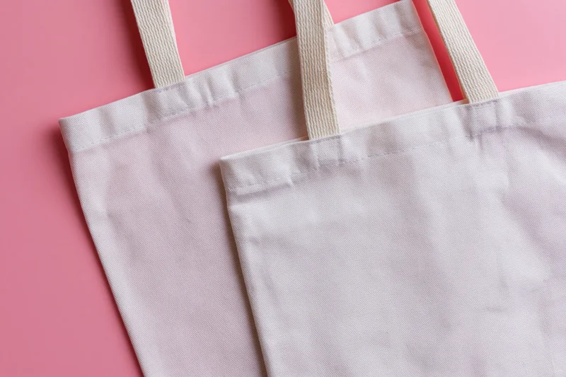 Two blank white canvas tote bags with woven cotton handles on pink background