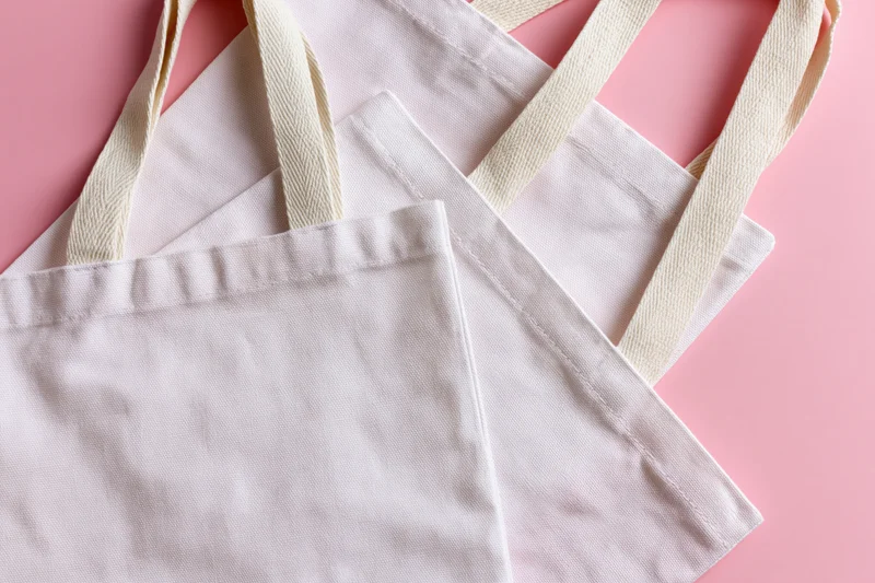 Two blank white canvas tote bags with woven cotton handles arranged on a pink surface