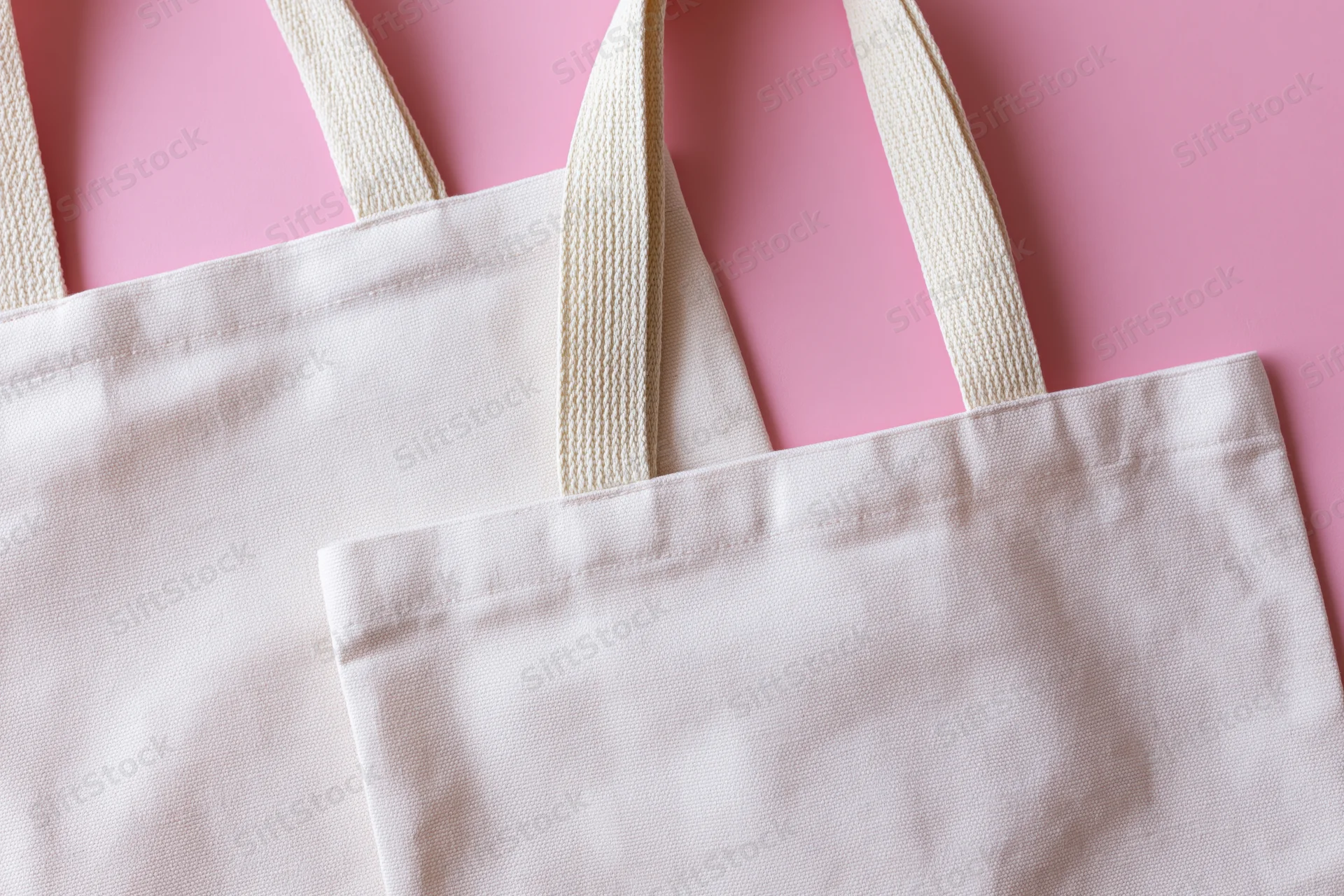 Two blank white canvas tote bags with woven cotton handles on pink background
