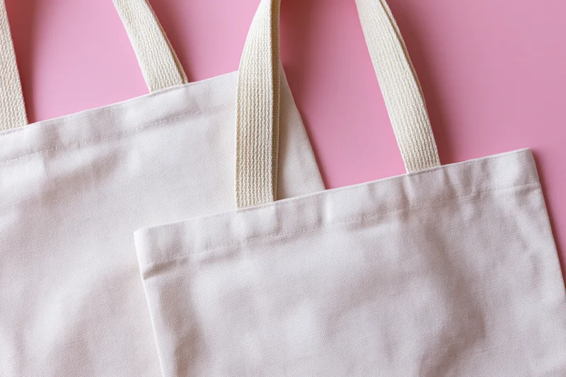 Two blank white canvas tote bags with woven cotton handles on pink background