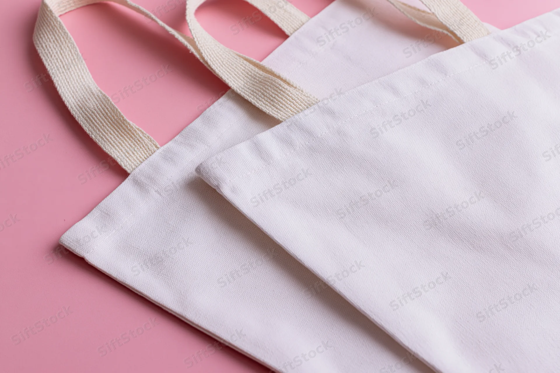 Two blank white canvas tote bags with woven cotton handles on pink surface