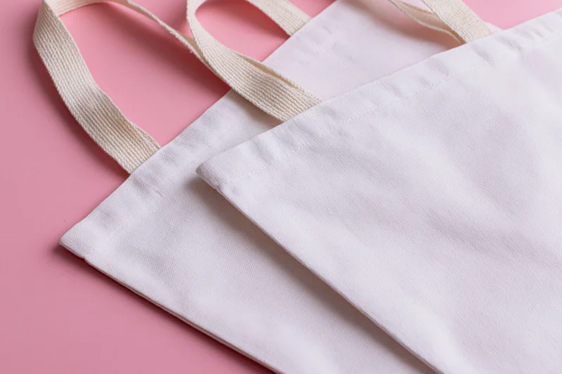 Two blank white canvas tote bags with woven cotton handles on pink surface