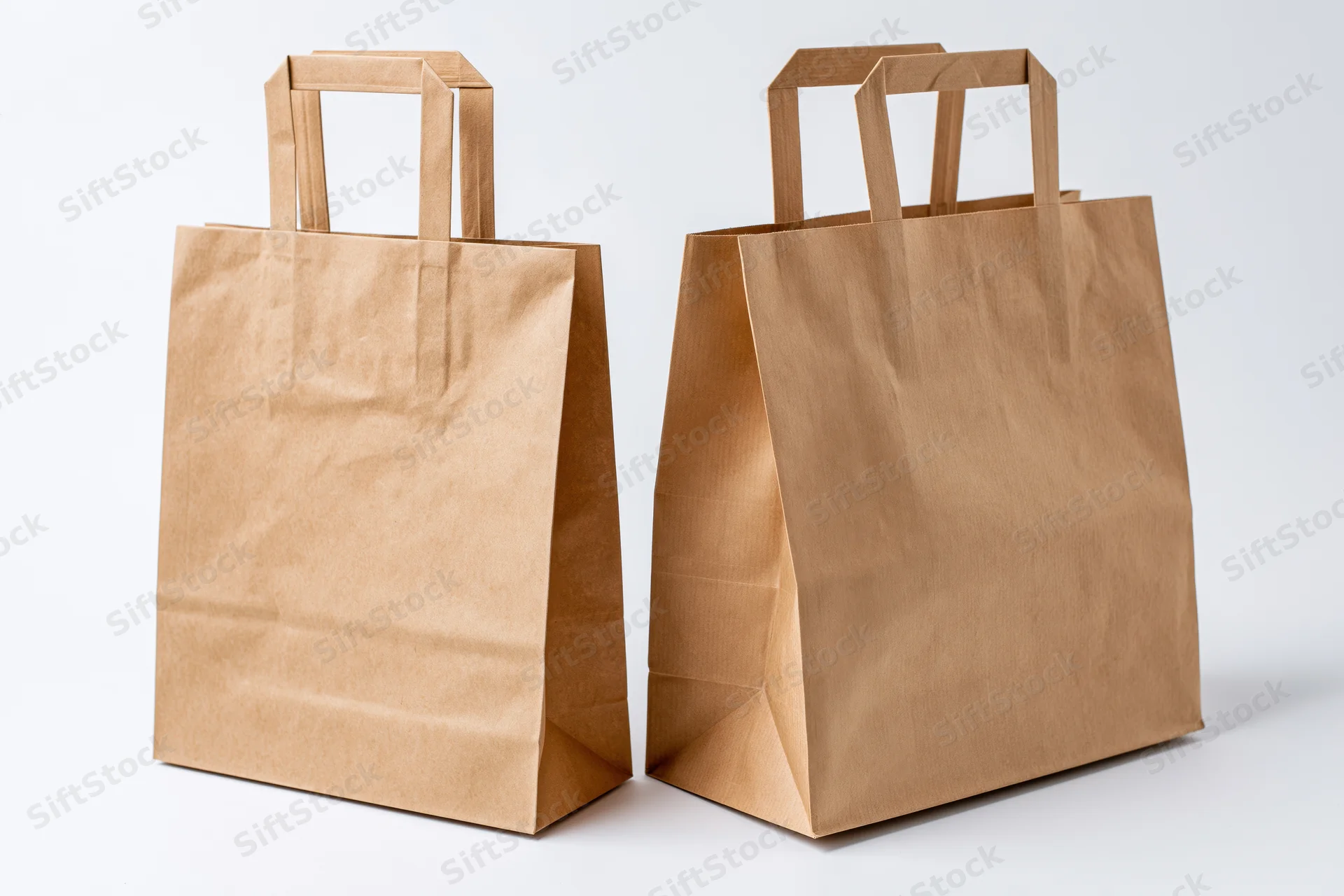 Two brown kraft paper shopping bags with flat handles standing side by side on white surface