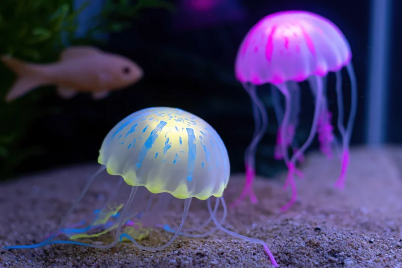 Two glowing silicone jellyfish decorations resting on sand in an aquarium with a fish in the background