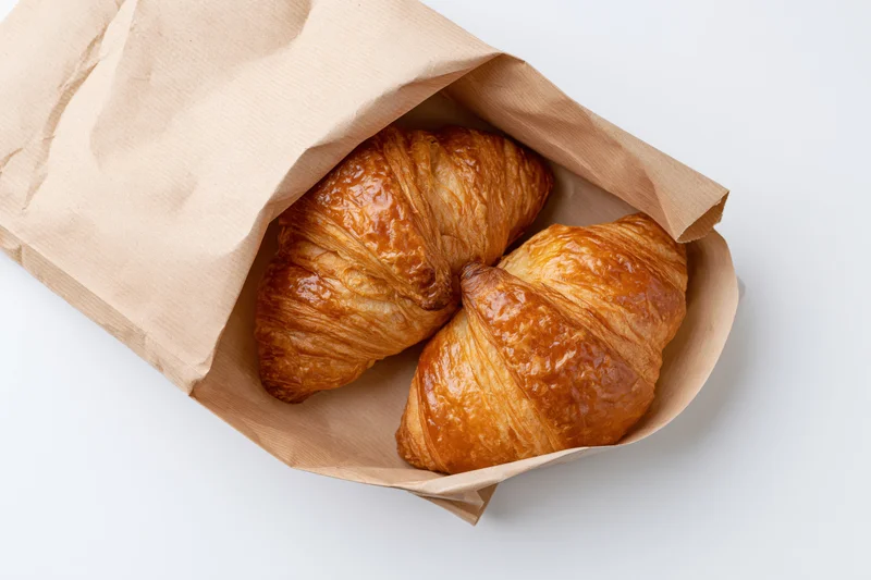 Two golden-brown croissants in an open kraft paper bag on white background