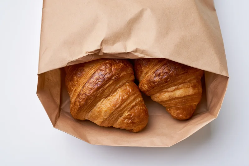 Two golden-brown croissants in an open kraft paper bag on white background
