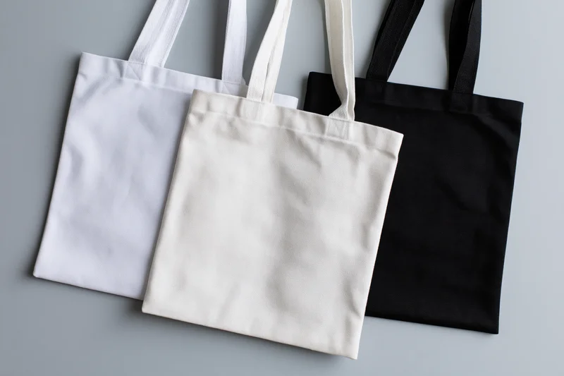 Three plain canvas tote bags in white, cream and black arranged flat on a gray surface