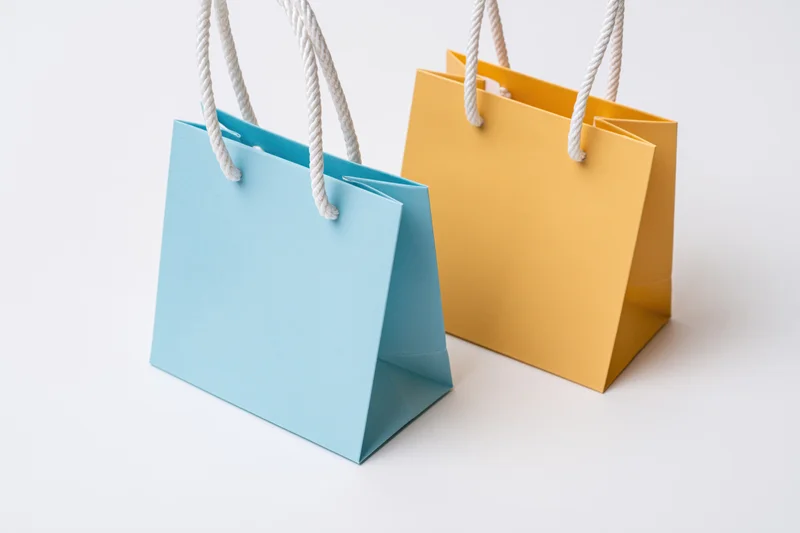 Two small paper shopping bags one light blue and one mustard on white surface