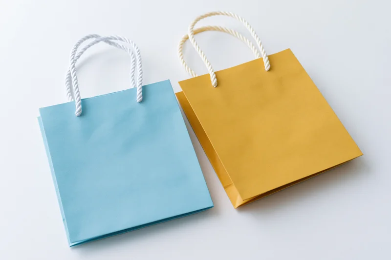 Two small paper shopping bags in light blue and mustard yellow with rope handles on white background
