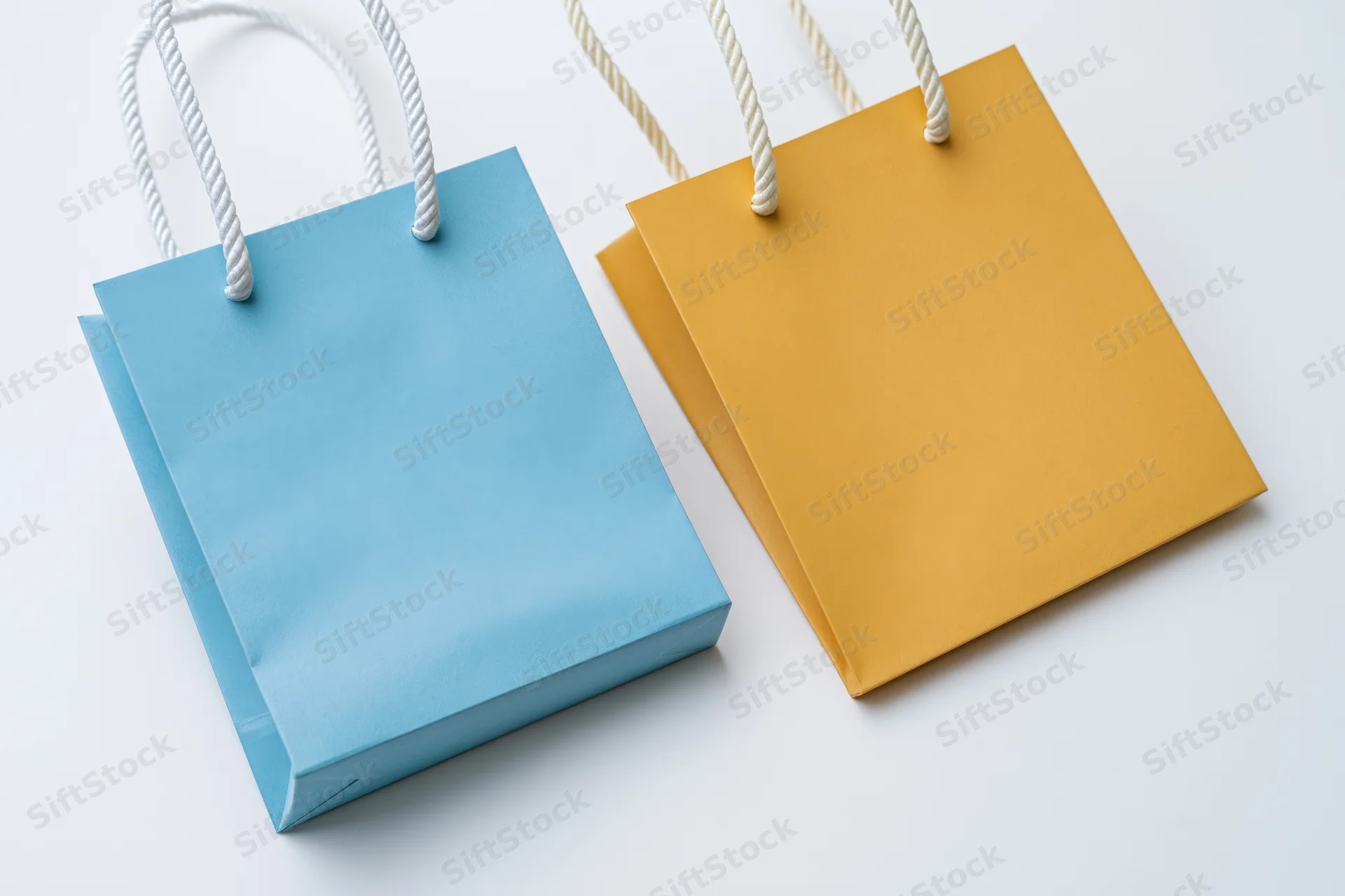 Two small paper shopping bags one light blue and one mustard on white surface