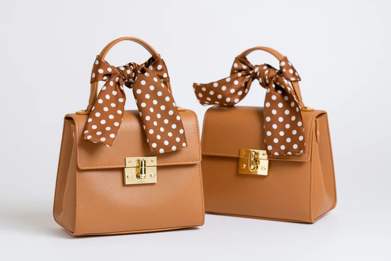 Two tan leather structured handbags with polka dot bows displayed side by side on white background