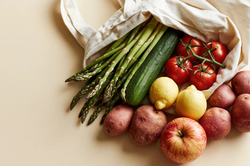 Open cotton tote bag spilling fresh vegetables and fruits onto a beige surface