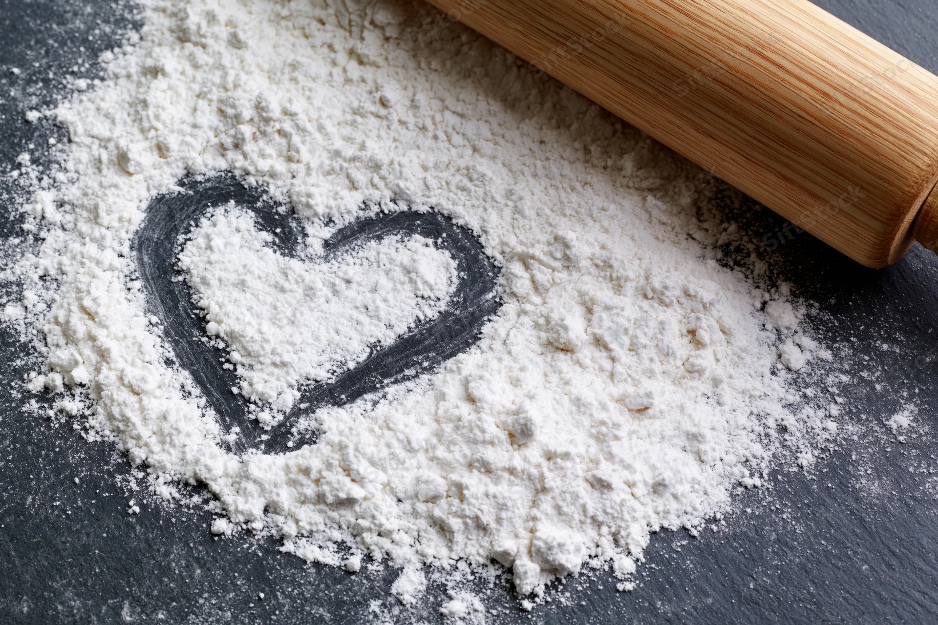Heart shaped in white flour on dark slate surface with wooden rolling pin nearby
