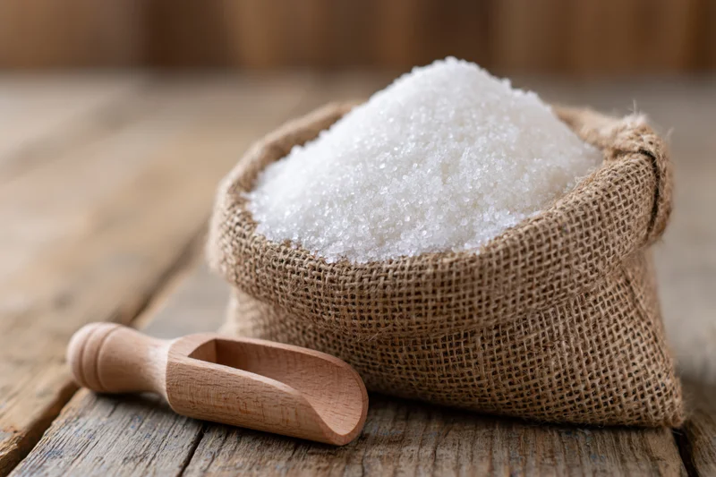 White granulated sugar piled in a small burlap sack with wooden scoop on rustic wooden table
